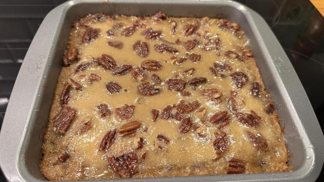 Le gâteau dans son moule gris carré. On voit les noix de pécan et un topping doré autour 
