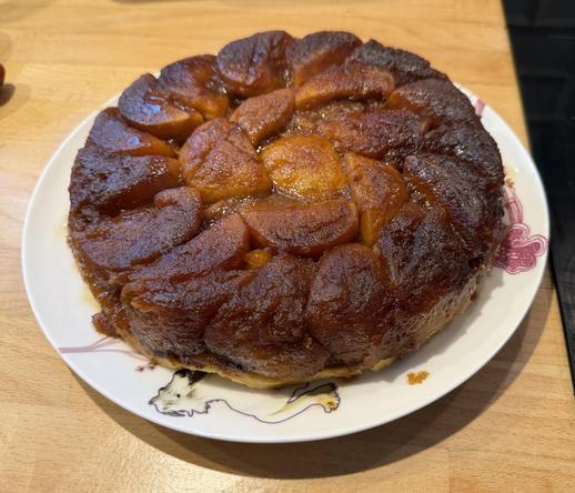 On voit la tarte tatin par-dessus sur une assiette blanche, colorée sur un plan de travail. On voit que les pommes sont un peu plus claires au centre, mais que le tout est vraiment très caramélisés et vraiment à l’air délicieux.