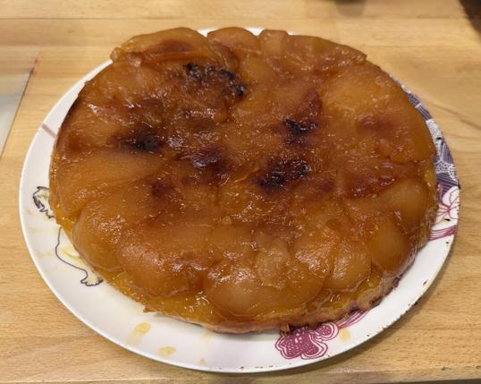 Une belle tarte tatin épaisse et très caramélisée sur une assiette blanche colorée sur un plan de travail en bois 
