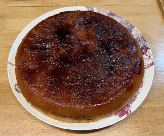 Une tarte Tatin très caramélisée. Elle a des zones bien foncées et est très belle et très lisse. Posee sur une grande assiette blanche avec des dessins, sur un plan de travail en hêtre clair. 