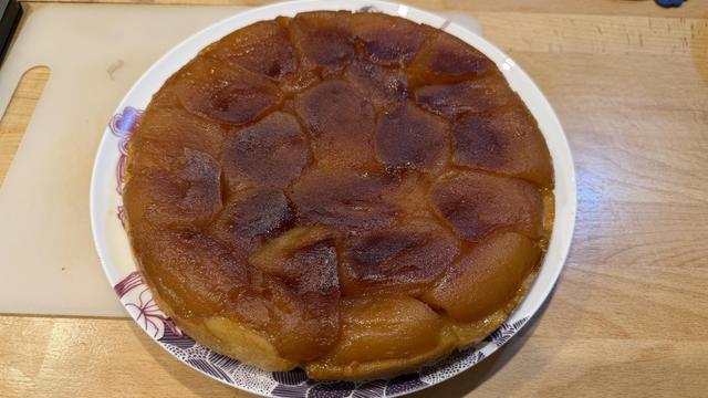 Une belle tarte Tatin avec le dessus bien caramélisé. On voit les pommes bien disposées et dorées marron. Sur une assiette blanche, sur un plan de travail en bois
