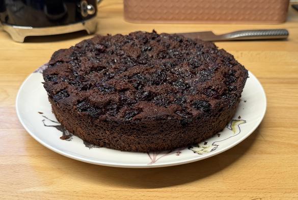 A round chocolate cake with a shiny top, decorated with dark fruit, is presented on a decorative plate. The background shows a wooden surface and kitchen utensils.