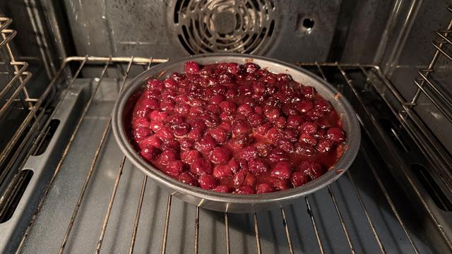 A metal baking dish filled with shiny, red cherries is placed inside an oven. The cherries are partially submerged in a glossy sauce, indicating they are being baked or cooked.