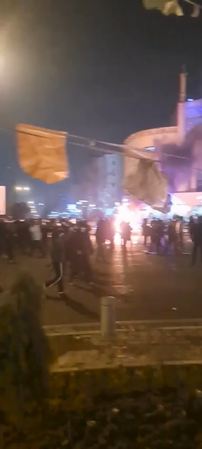 On voit un bâtiment en flammes avec plein de manifestants devant. Images floues prises la nuit. 