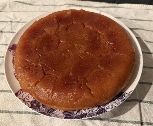 Une belle tarte tatin bien ronde et très caramélisée et foncée, posée sur une assiette blanche qui elle-même repose sur un torchon blanc avec des rayures vertes. Elle a l’air très appétissante.