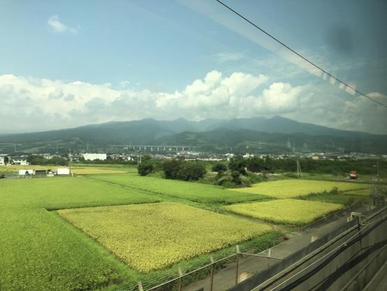 Photo prise de la fenêtre d’un train. On voit des champs très verts en premier plan et une petite montagne au loin avec de beaux nuages blancs 