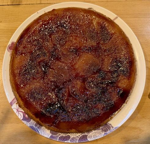 Une belle tarte, tatin, très caramélisée, avec les pommes aplaties au-dessus. La vue est prise d’eau au-dessus, on voit un plan de travail en bois autour et des reflets sur les pommes.