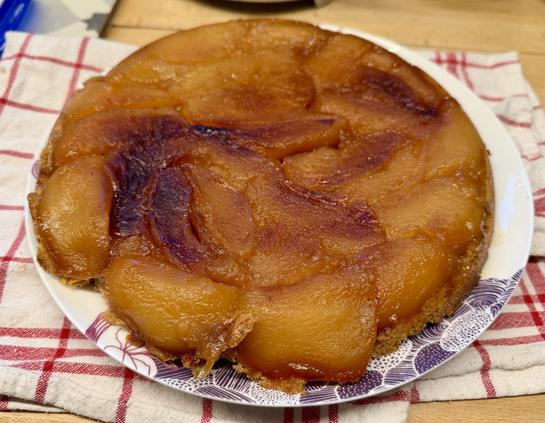 Une superbe tarte, tatin, très dorée et caramélisées sur une assiette qui a des dessins violet, qui elle-même est posé sur un torchon rouge et blanc. Le tout est extrêmement appétissant.